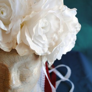 May include: A headpiece featuring a cluster of white fabric roses with a subtle glitter finish, resting atop a mannequin head covered in vintage-style printed paper. The piece includes a white lace trim and a deep red ribbon hanging down the side.