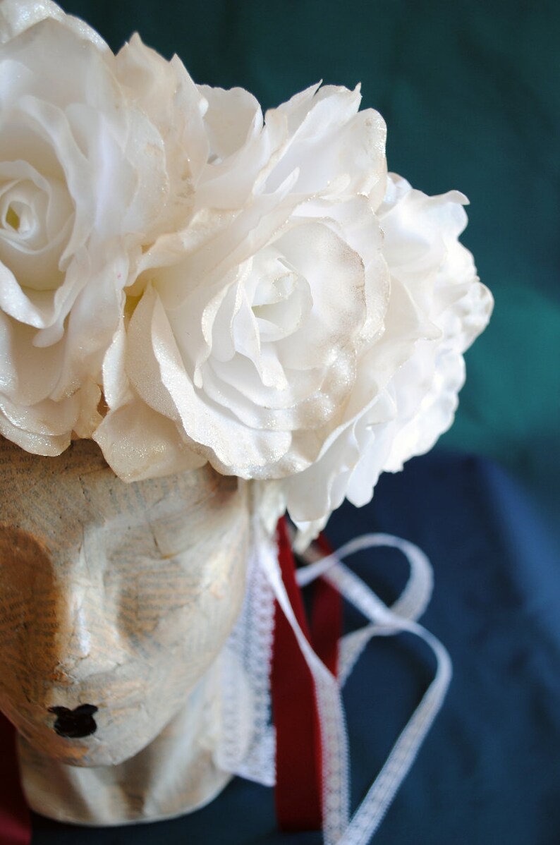 May include: A headpiece featuring a cluster of white fabric roses with a subtle glitter finish, resting atop a mannequin head covered in vintage-style printed paper. The piece includes a white lace trim and a deep red ribbon hanging down the side.