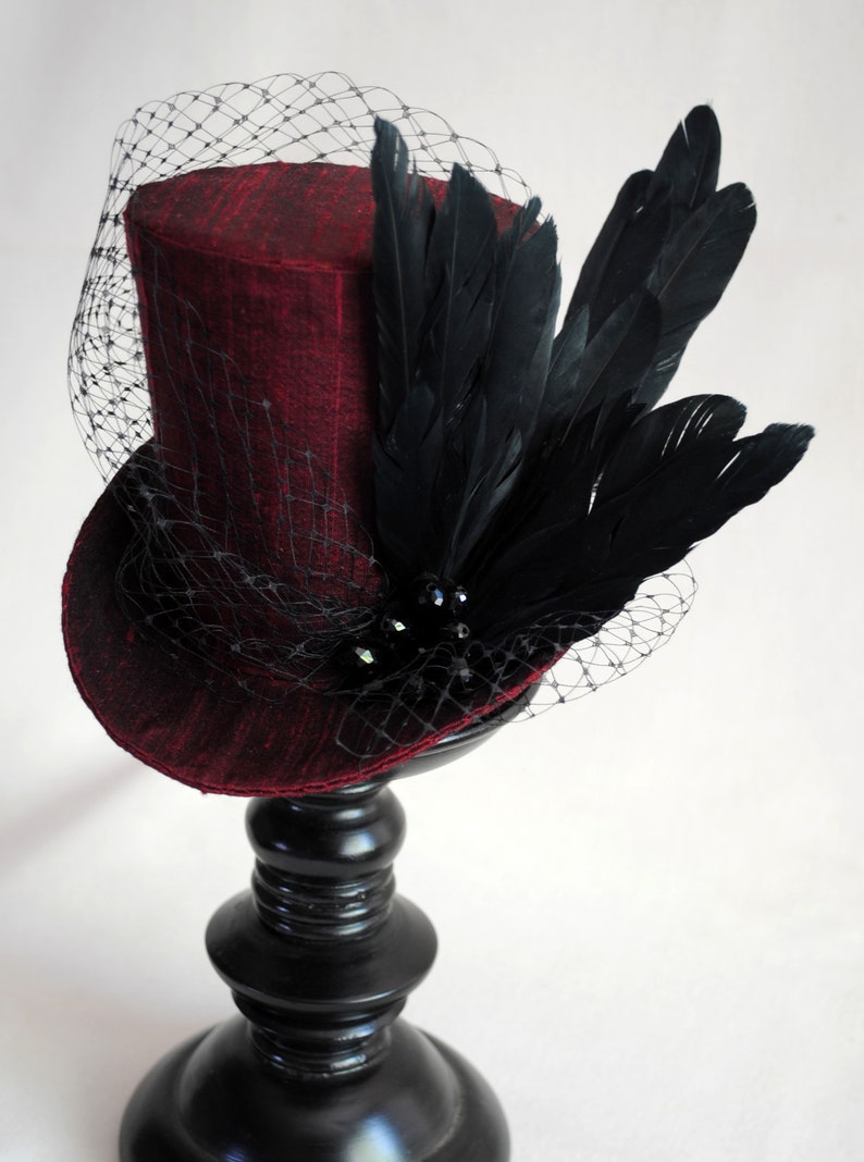 May include: A deep red top hat featuring a textured fabric finish, adorned with a cluster of black feathers and black faceted beads. A fine black netting veil extends from the crown over the brim, creating a layered look on the structured hat.