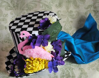 May include: A top hat featuring a black and white checkered pattern, adorned with a pink felt flamingo, vibrant yellow chrysanthemum-style flowers, deep purple hydrangea blossoms, green fabric leaves, and a flowing bright blue fabric sash.
