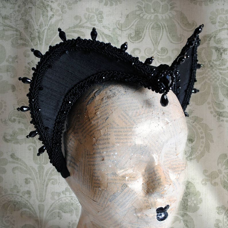 May include: A black headpiece featuring a structured, arched frame adorned with intricate braided trim and dangling black teardrop beads along the outer edge. A large, faceted black stone sits at the center, surrounded by smaller black beads.