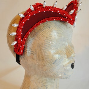May include: A deep red fabric headpiece featuring a curved, elevated structure adorned with intricate red braided trim and numerous white teardrop-shaped faux pearls lining the edges. The headpiece is secured with a black elastic band.