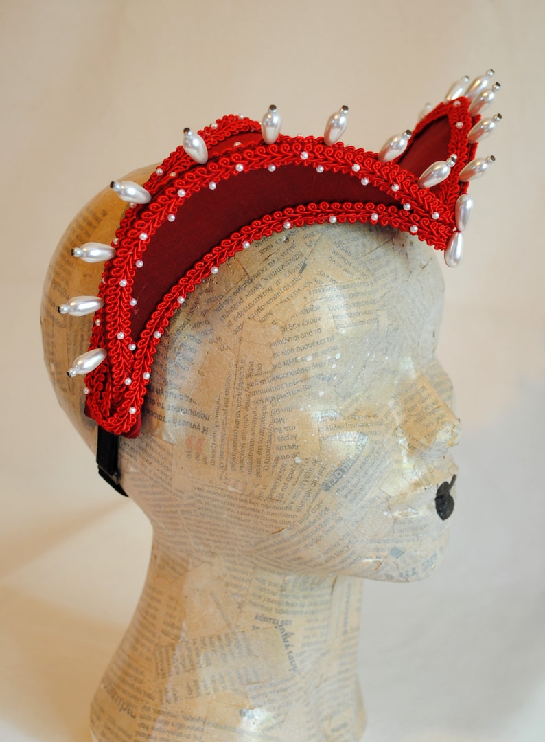 May include: A deep red fabric headpiece featuring a curved, elevated structure adorned with intricate red braided trim and numerous white teardrop-shaped faux pearls lining the edges. The headpiece is secured with a black elastic band.