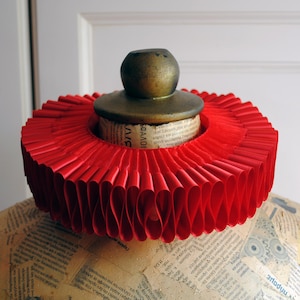 May include: A vibrant red ruffled collar, likely for a costume or historical reenactment, is displayed on a mannequin. The collar is made of pleated fabric and features a decorative brass-colored top. The mannequin's form is covered in aged paper.
