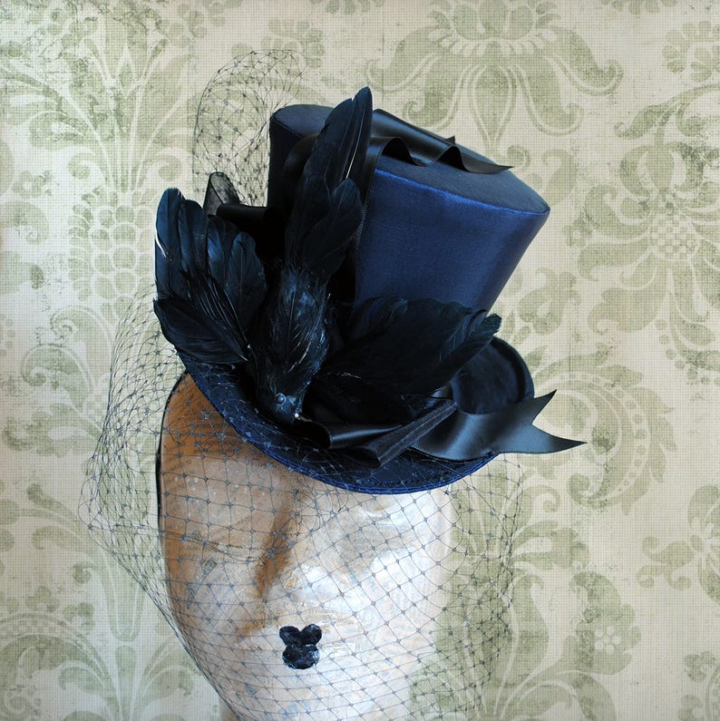 May include: A blue top hat with a black velvet band and black feathers. The hat is on a mannequin head with a black net veil.