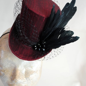 May include: A deep red top hat fascinator featuring a textured fabric finish, adorned with a black netting veil, a cluster of dark beads, and a spray of black feathers. The hat is secured with a thin black headband for placement on the head.