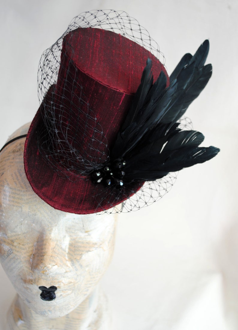 May include: A deep red top hat fascinator featuring a textured fabric finish, adorned with a black netting veil, a cluster of dark beads, and a spray of black feathers. The hat is secured with a thin black headband for placement on the head.