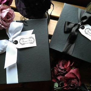 May include: Two square black gift boxes, one tied with a white ribbon and the other with a black ribbon. Each box features a white rectangular tag with the text Bizane Noir in a decorative script font, attached by a black string to the ribbon.