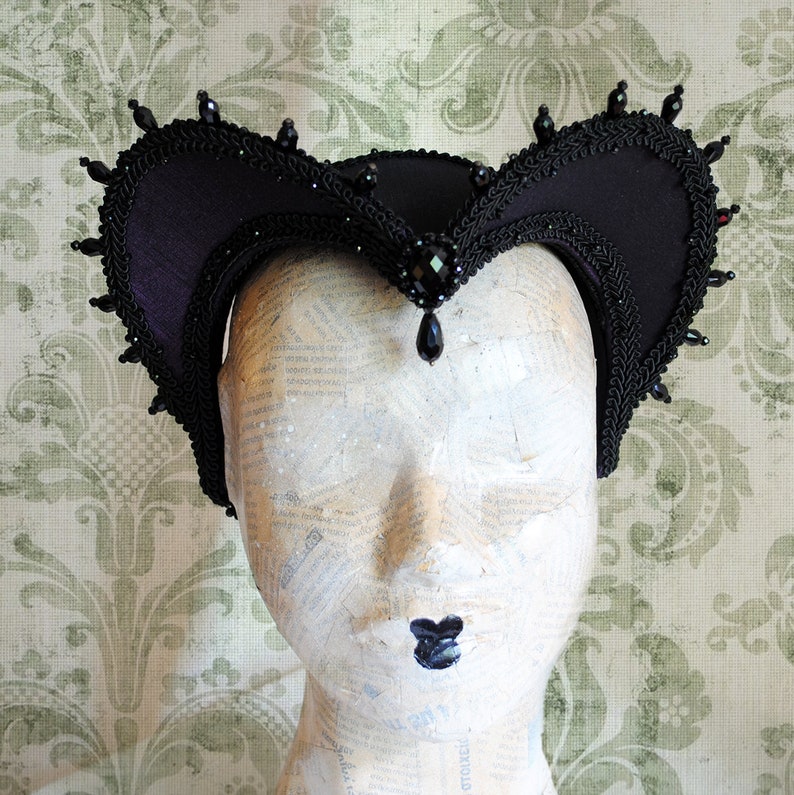 May include: A black velvet headband with a pointed crown shape, adorned with black beads and a dangling black gemstone.