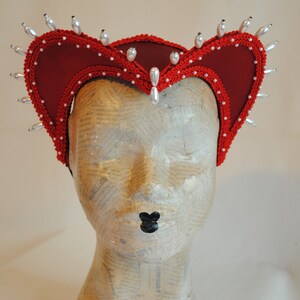 May include: A deep red fabric headpiece featuring a structured, heart-shaped silhouette with a prominent central peak. The edges are trimmed with intricate red braided cord and adorned with numerous white, teardrop-shaped faux pearls.
