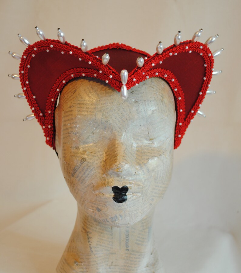 May include: A deep red fabric headpiece featuring a structured, heart-shaped silhouette with a prominent central peak. The edges are trimmed with intricate red braided cord and adorned with numerous white, teardrop-shaped faux pearls.