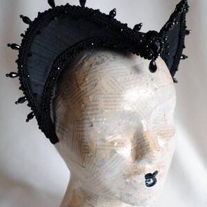 May include: A black headpiece featuring a high, arched structure with intricate braided trim and dangling black beads along the top edge. The piece is adorned with a central faceted black stone and smaller bead accents, creating a structured, crown-like appearance.