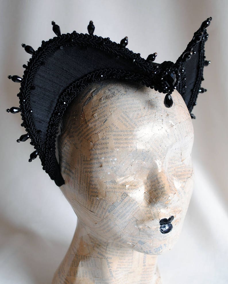 May include: A black headpiece featuring a high, arched structure with intricate braided trim and dangling black beads along the top edge. The piece is adorned with a central faceted black stone and smaller bead accents, creating a structured, crown-like appearance.