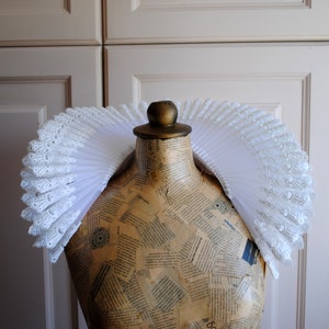 May include: A white lace and tulle ruffled collar, also known as a ruff, on a dress form. The ruff is a large, circular collar that is often worn as a costume accessory.