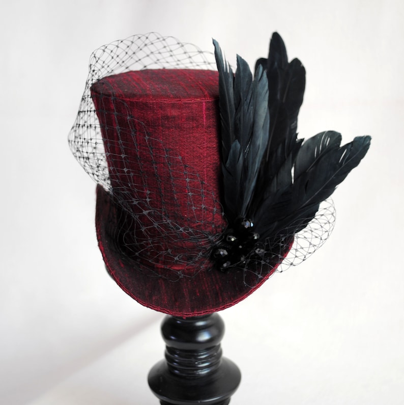 May include: A deep red top hat featuring a textured fabric finish, adorned with a cluster of black feathers and dark beads. A fine black netting veil extends from the side, draping over the crown and brim of the structured cylindrical hat.