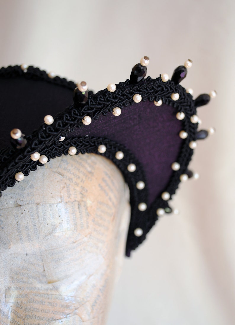 May include: A black headband with a purple fabric base and a black lace trim. The headband is decorated with white pearls and black beads.