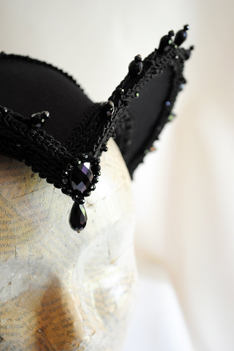 May include: A black lace headband with a large black gemstone pendant. The headband is decorated with black beads and has a pointed shape.