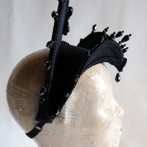 May include: A black fabric headpiece with a pointed crown and a curved brim. The headpiece is adorned with black beads and is worn on a mannequin head.