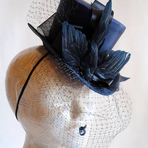 May include: A navy blue top hat with a black ribbon and black feathers, adorned with a black veil.