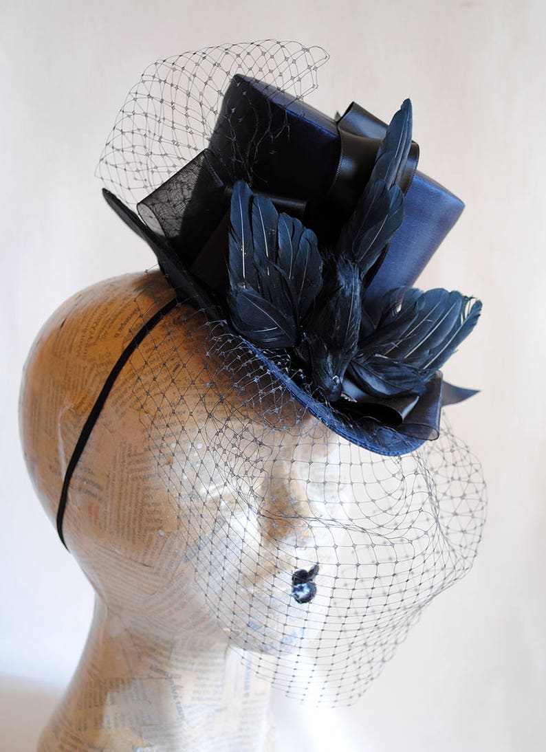 May include: A navy blue top hat with a black ribbon and black feathers, adorned with a black veil.