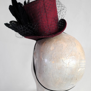 May include: A deep burgundy mini top hat fascinator featuring a structured cylindrical crown and a curved brim. The hat is adorned with a cluster of black feathers and a fine black netting veil that drapes over the front and side of the hat.
