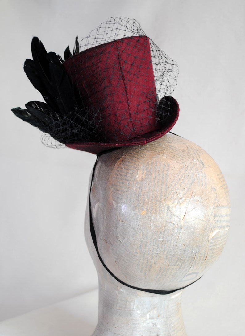 May include: A deep burgundy mini top hat fascinator featuring a structured cylindrical crown and a curved brim. The hat is adorned with a cluster of black feathers and a fine black netting veil that drapes over the front and side of the hat.
