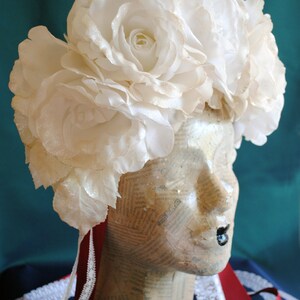 May include: A floral headpiece featuring large, white, shimmering fabric roses arranged in a crown. The headpiece is displayed on a mannequin head covered in vintage newspaper print, accented with long, dark red ribbons and white lace trim.