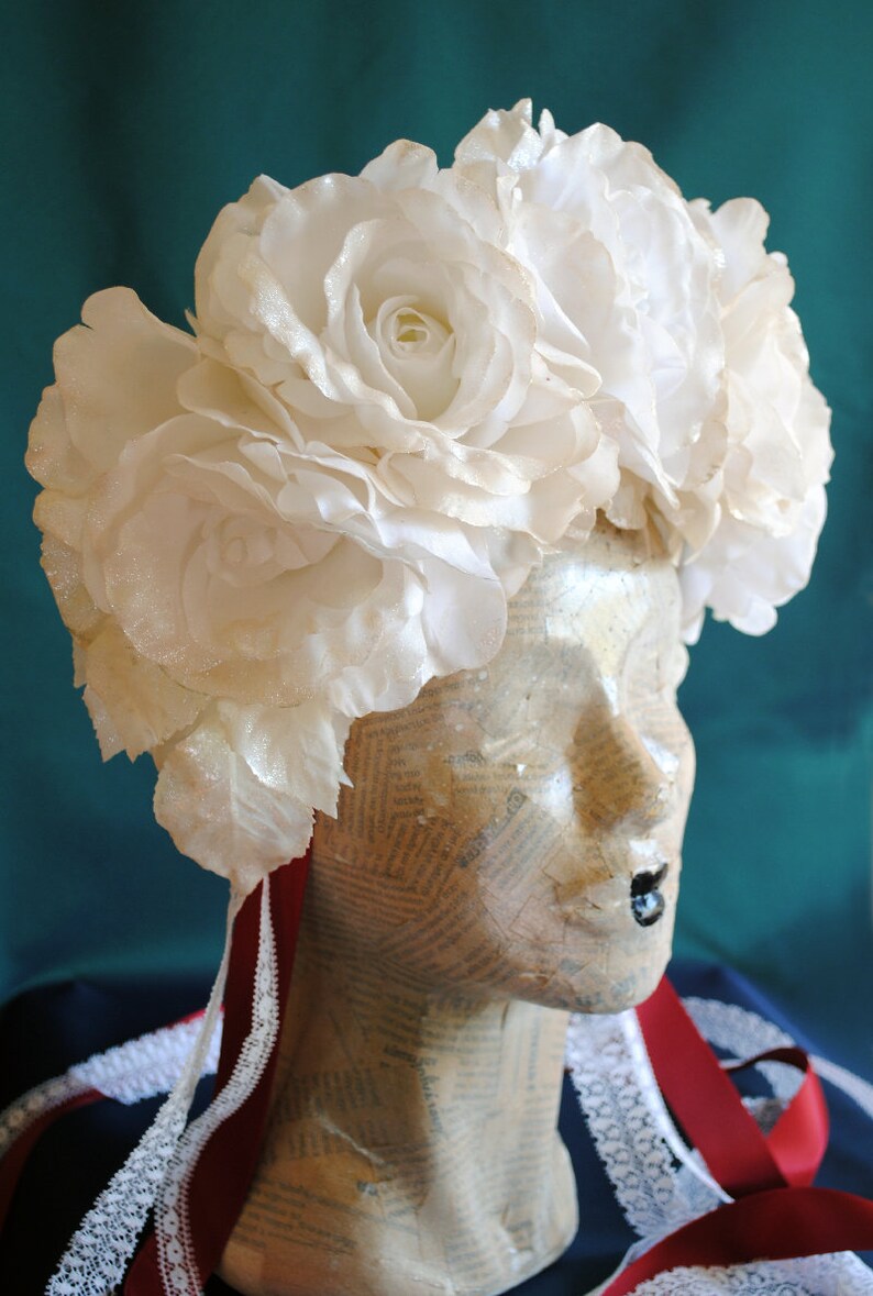 May include: A floral headpiece featuring large, white, shimmering fabric roses arranged in a crown. The headpiece is displayed on a mannequin head covered in vintage newspaper print, accented with long, dark red ribbons and white lace trim.