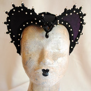 May include: A black felt headpiece with a pointed crown and curved sides. The headpiece is decorated with black and white beads and a large black gemstone.