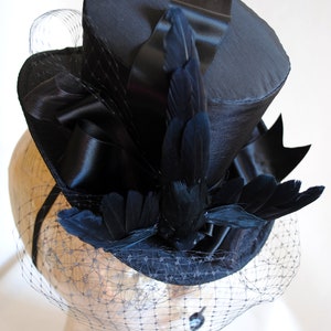 May include: A black top hat with a black satin ribbon and black feathers. The hat is adorned with a black veil.