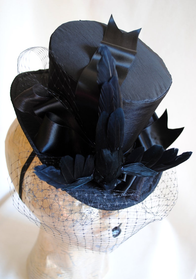 May include: A black top hat with a black satin ribbon and black feathers. The hat is adorned with a black veil.