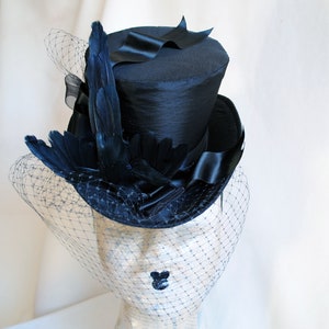 May include: A black top hat with a black feather and veil. The hat is made of satin and has a tall, cylindrical crown.