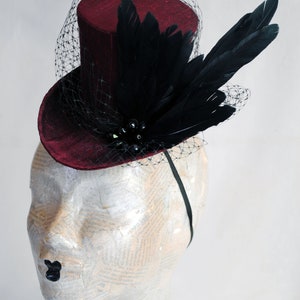 May include: A deep burgundy mini top hat featuring a black netting veil, a cluster of black feathers, and dark beads. The hat is secured to a thin black headband, creating a structured headpiece with a textured fabric finish and dark accents.