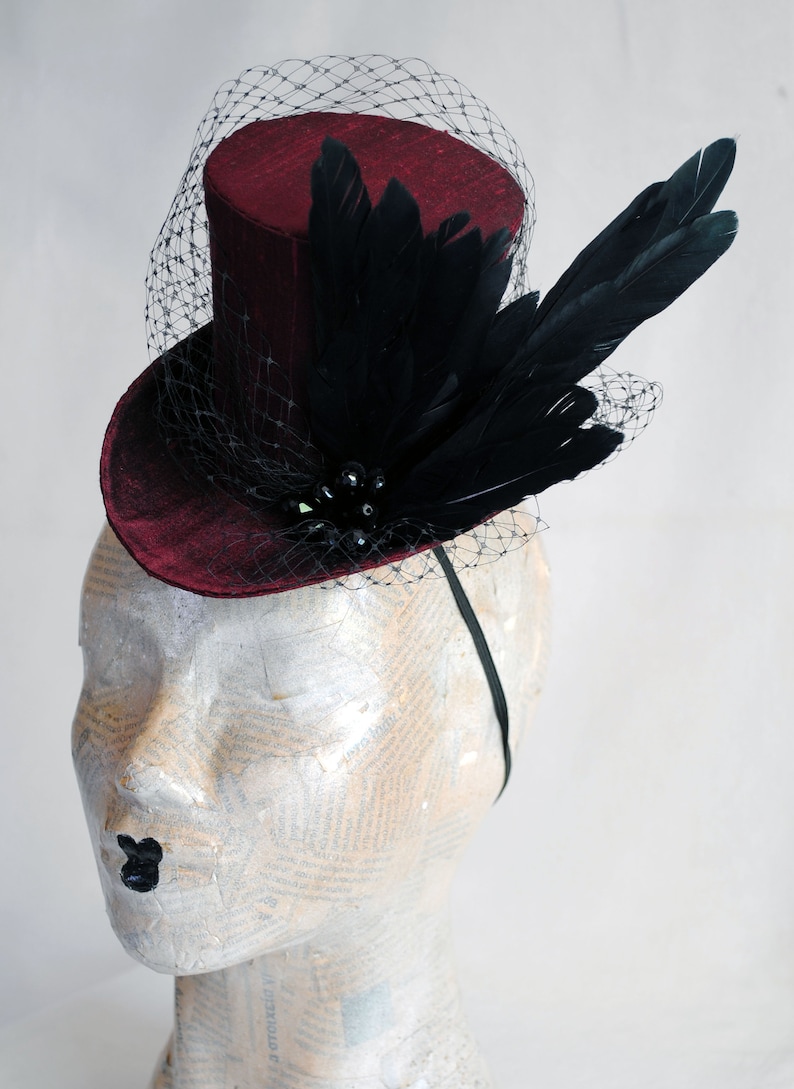 May include: A deep burgundy mini top hat featuring a black netting veil, a cluster of black feathers, and dark beads. The hat is secured to a thin black headband, creating a structured headpiece with a textured fabric finish and dark accents.