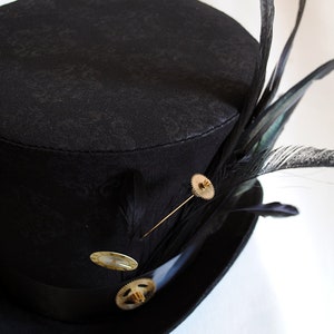 May include: A black top hat with a patterned design, adorned with black feathers and gold-colored embellishments. The hat features a black band and is decorated with a gear-shaped pin and other decorative pins. A stylish accessory.