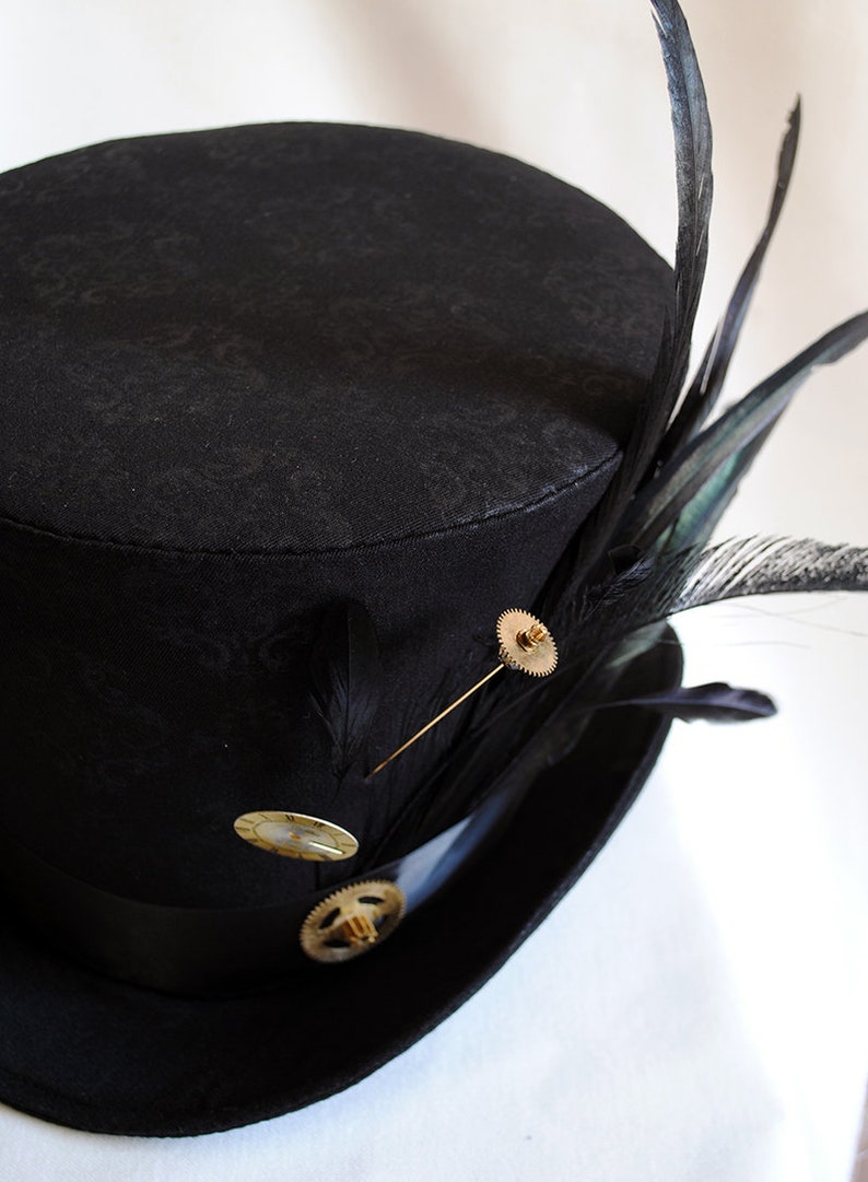 May include: A black top hat with a patterned design, adorned with black feathers and gold-colored embellishments. The hat features a black band and is decorated with a gear-shaped pin and other decorative pins. A stylish accessory.