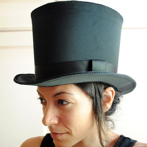 May include: A woman wearing a black top hat with a black band. The hat is tall and has a wide brim. The woman has dark hair and is looking down.