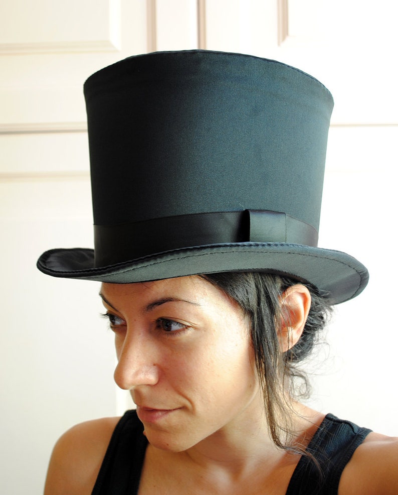 May include: A woman wearing a black top hat with a black band. The hat is tall and has a wide brim. The woman has dark hair and is looking down.