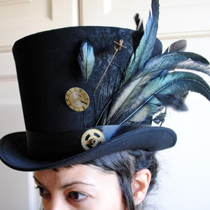 May include: Black top hat with a decorative design, adorned with feathers, a clock face, and a gear. The hat features a black band and is embellished with a gold-colored pin. The feathers have shades of blue and green.
