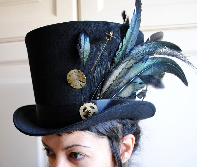 May include: Black top hat with a decorative design, adorned with feathers, a clock face, and a gear. The hat features a black band and is embellished with a gold-colored pin. The feathers have shades of blue and green.