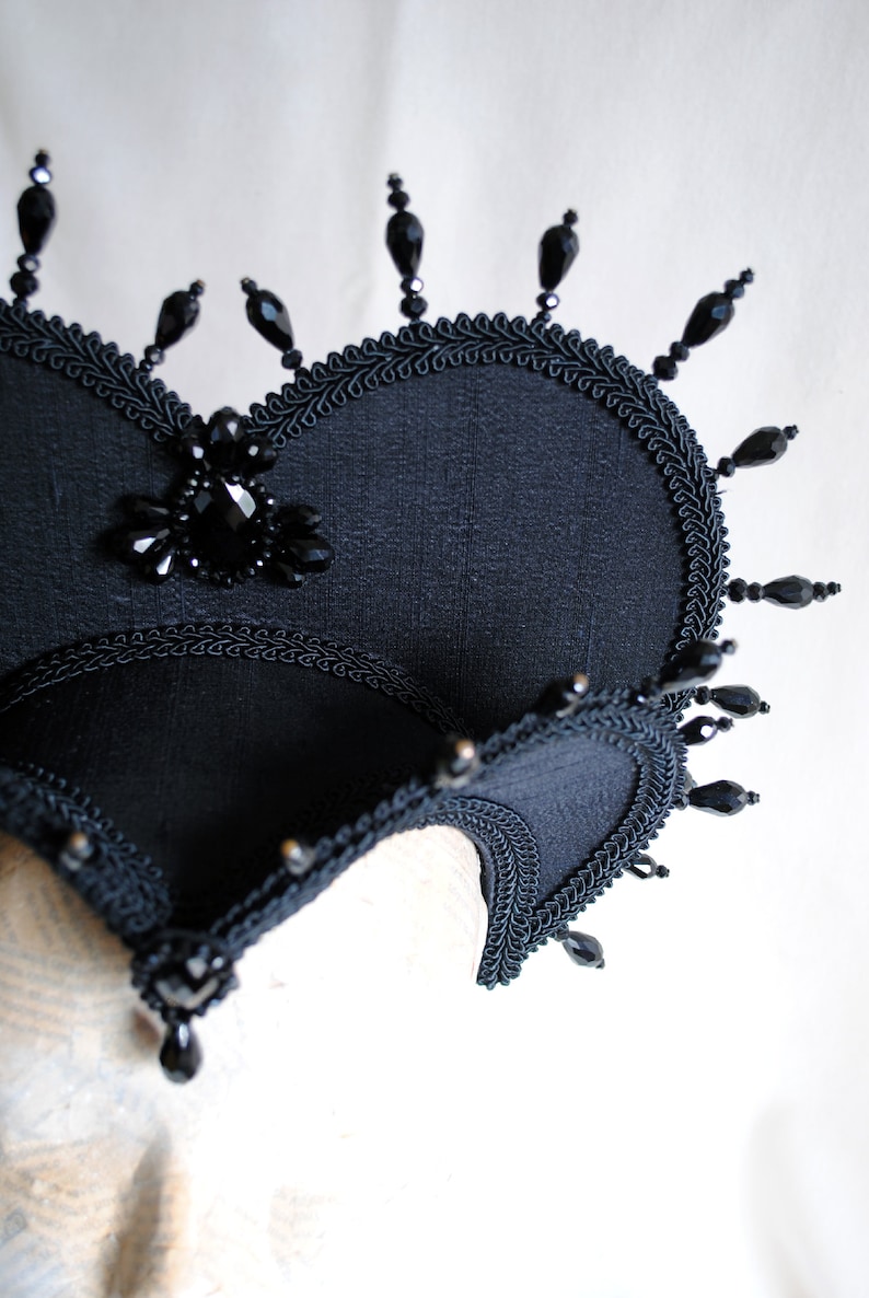 May include: A black fabric crown with a large black jewel in the center and black beads around the edges.