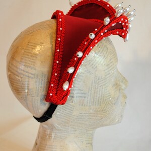 May include: A deep red fabric headpiece featuring a structured, elevated design adorned with intricate red braided trim and numerous white pearl-like beads. The accessory is secured with a black elastic strap and displays a crown-like silhouette.