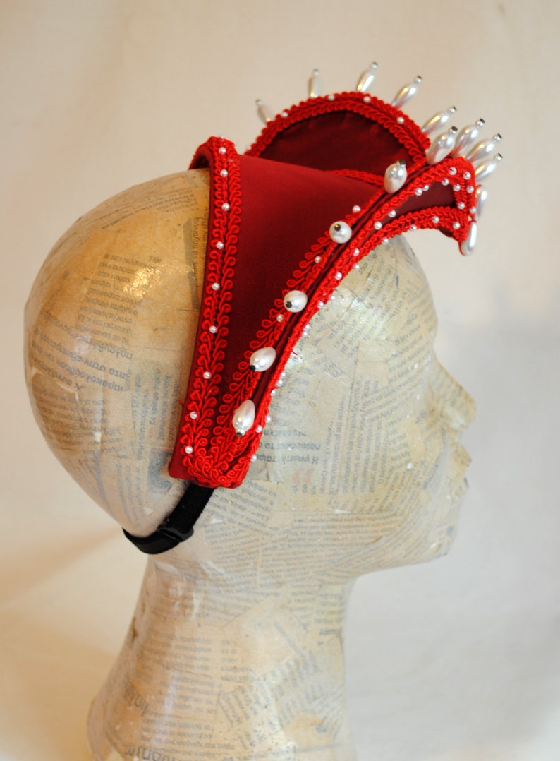 May include: A deep red fabric headpiece featuring a structured, elevated design adorned with intricate red braided trim and numerous white pearl-like beads. The accessory is secured with a black elastic strap and displays a crown-like silhouette.