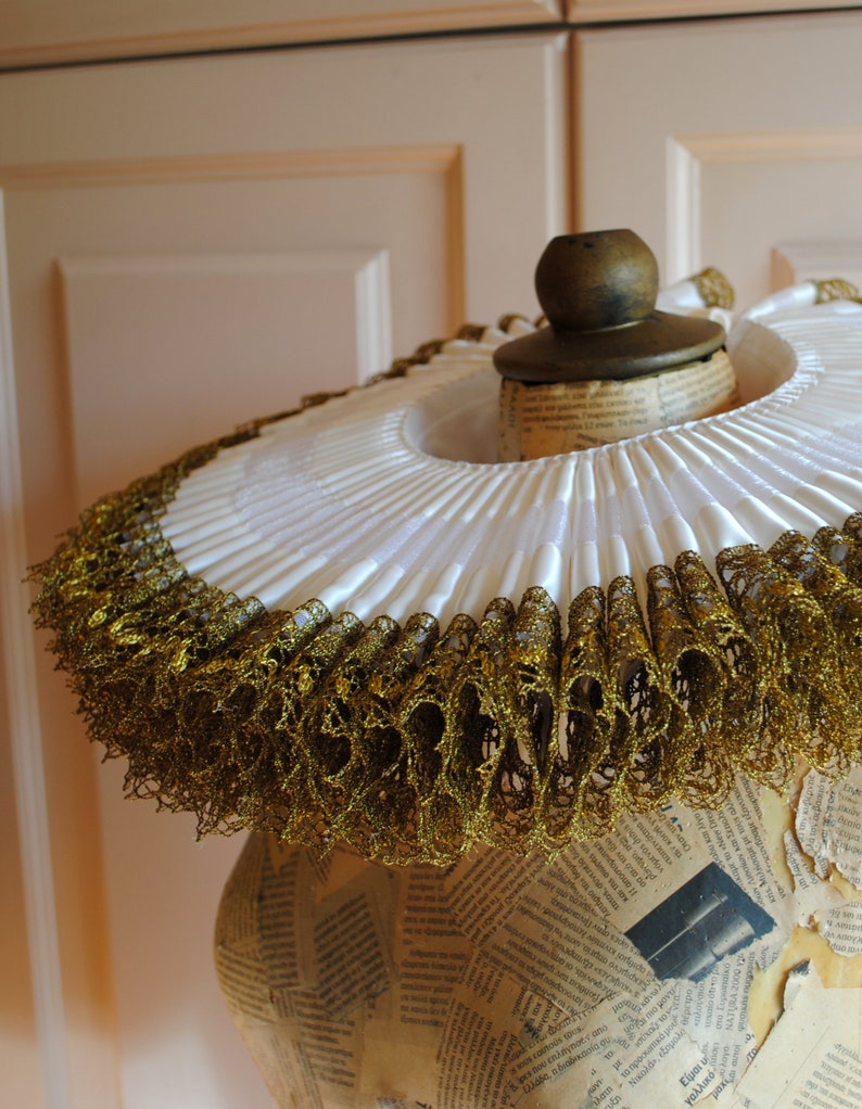 K&ouml;nnte beinhalten: Ein wei&szlig;er R&uuml;schenkragen mit goldener Spitzenborte. Der Kragen befindet sich auf einer mit Zeitungspapier bedeckten Schaufensterpuppe.