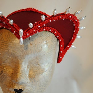 May include: A deep red fabric headpiece featuring a structured, curved brim edged with intricate red braided trim. The headpiece is adorned with numerous small white pearls and several larger, teardrop-shaped white faux pearls along the outer edge.