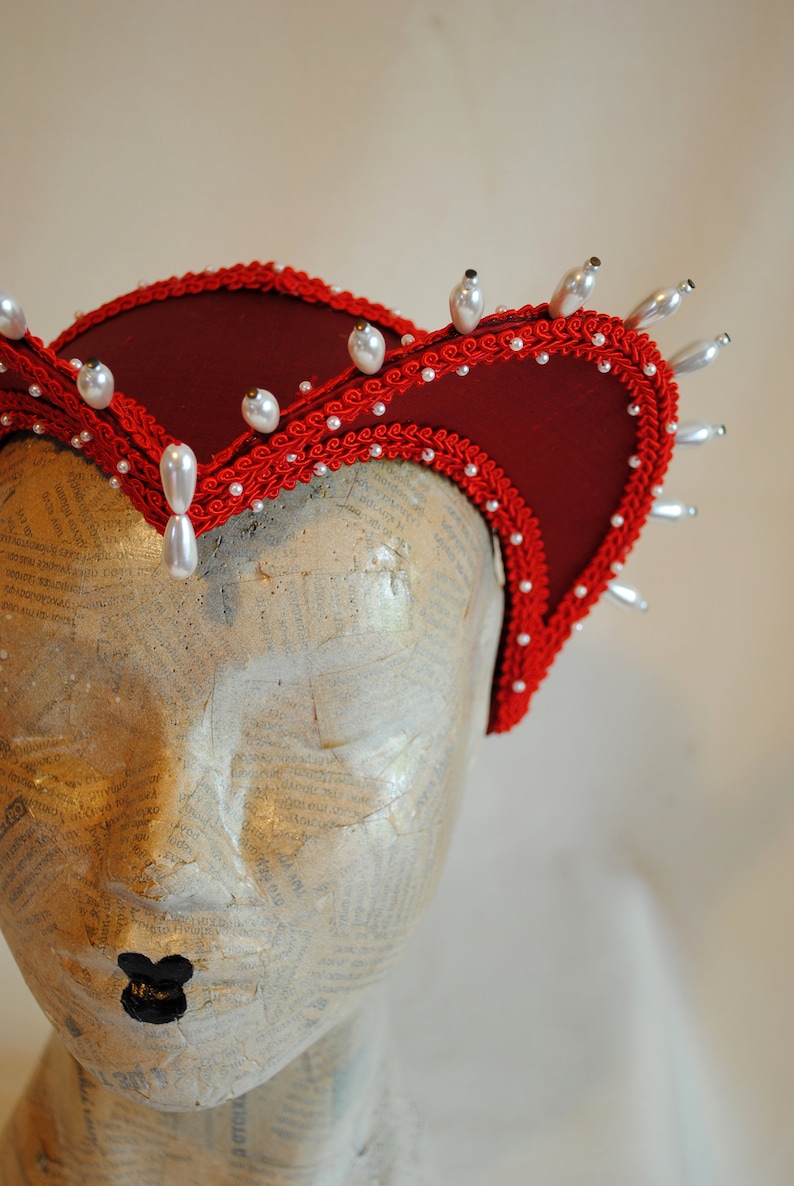 May include: A deep red fabric headpiece featuring a structured, curved brim edged with intricate red braided trim. The headpiece is adorned with numerous small white pearls and several larger, teardrop-shaped white faux pearls along the outer edge.