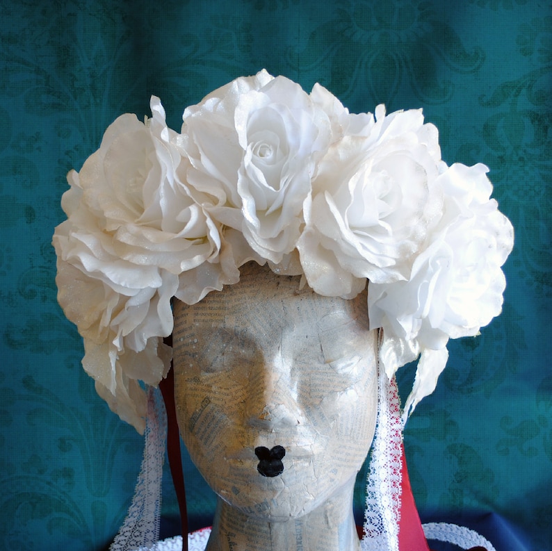 May include: A floral headpiece featuring a dense arrangement of large, white fabric roses. The crown is displayed on a mannequin head and includes two long, thin red ribbons and white lace trim hanging down from the sides of the floral arrangement.