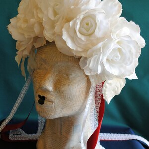 May include: A floral headpiece featuring large white roses with a subtle shimmer, worn on a mannequin head covered in printed text. The headpiece includes long, trailing white lace ribbons and solid red satin ribbons hanging down the sides.