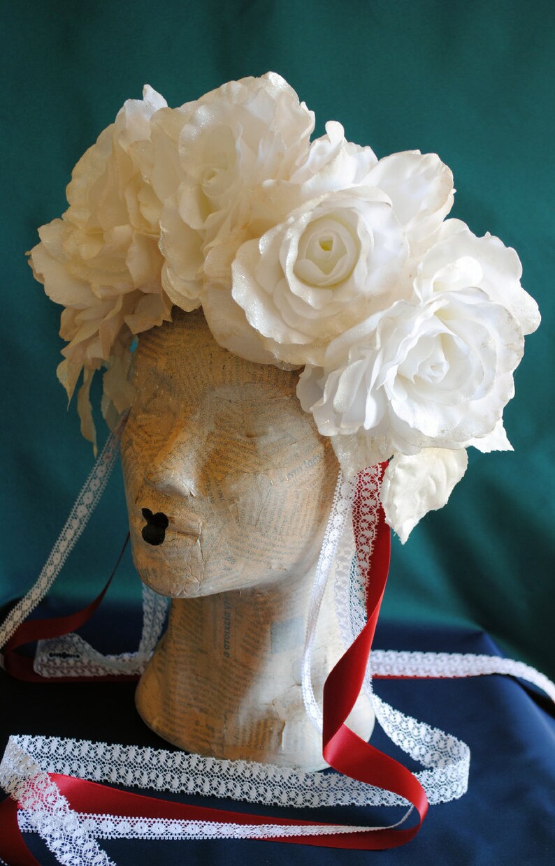 May include: A floral headpiece featuring large white roses with a subtle shimmer, worn on a mannequin head covered in printed text. The headpiece includes long, trailing white lace ribbons and solid red satin ribbons hanging down the sides.
