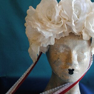 May include: A headpiece featuring a cluster of large, white fabric roses with a subtle shimmer. The floral arrangement is attached to a mannequin head covered in newsprint, with two long, dark red velvet ribbons trimmed in white lace hanging down.
